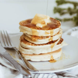 Maple syrup being poured over a stack of fluffy pancakes topped with a melting pat of butter