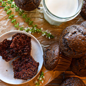 Freshly baked chocolate muffins on a cooling rack next to a glass of milk and a broken muffin showing its moist crumb