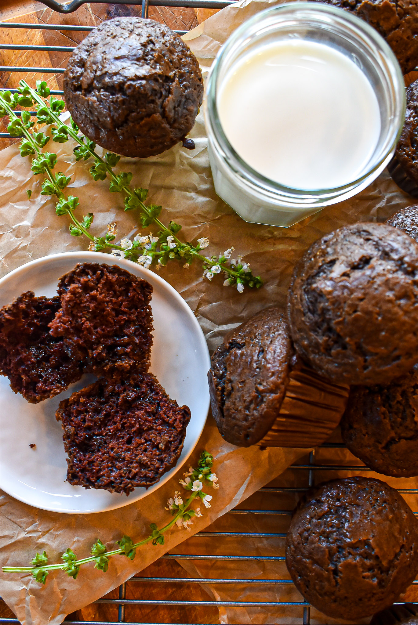 Freshly baked chocolate muffins on a cooling rack next to a glass of milk and a broken muffin showing its moist crumb