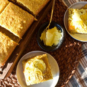 Golden cornbread squares on a wooden tray, served alongside individual slices topped with butter and honey on small plates
