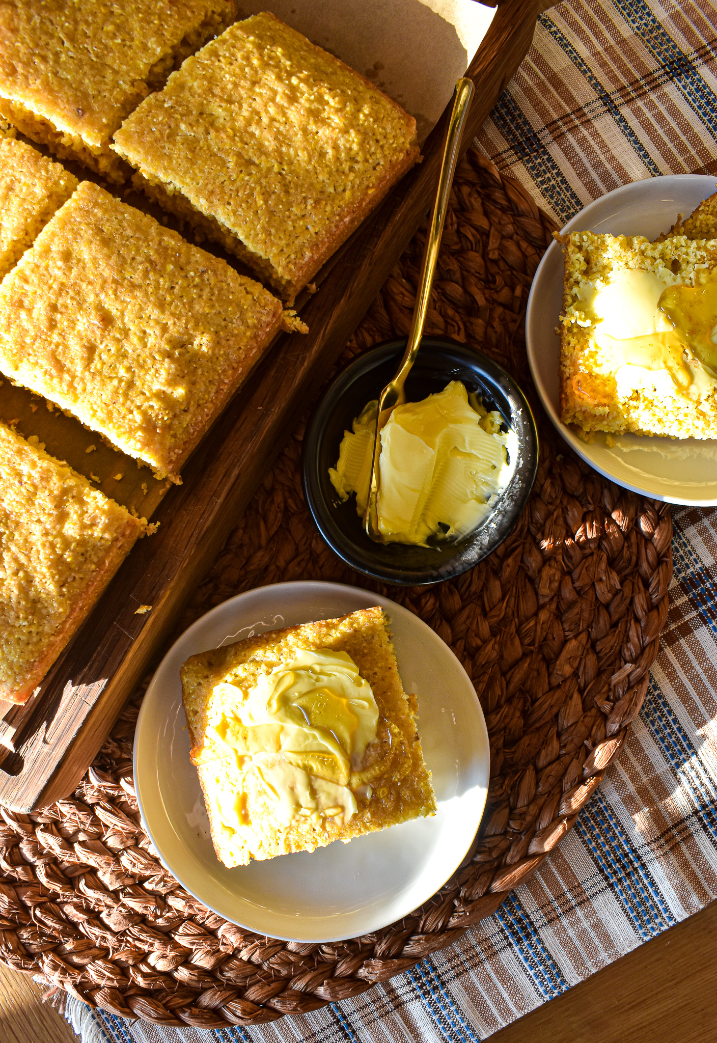 Golden cornbread squares on a wooden tray, served alongside individual slices topped with butter and honey on small plates
