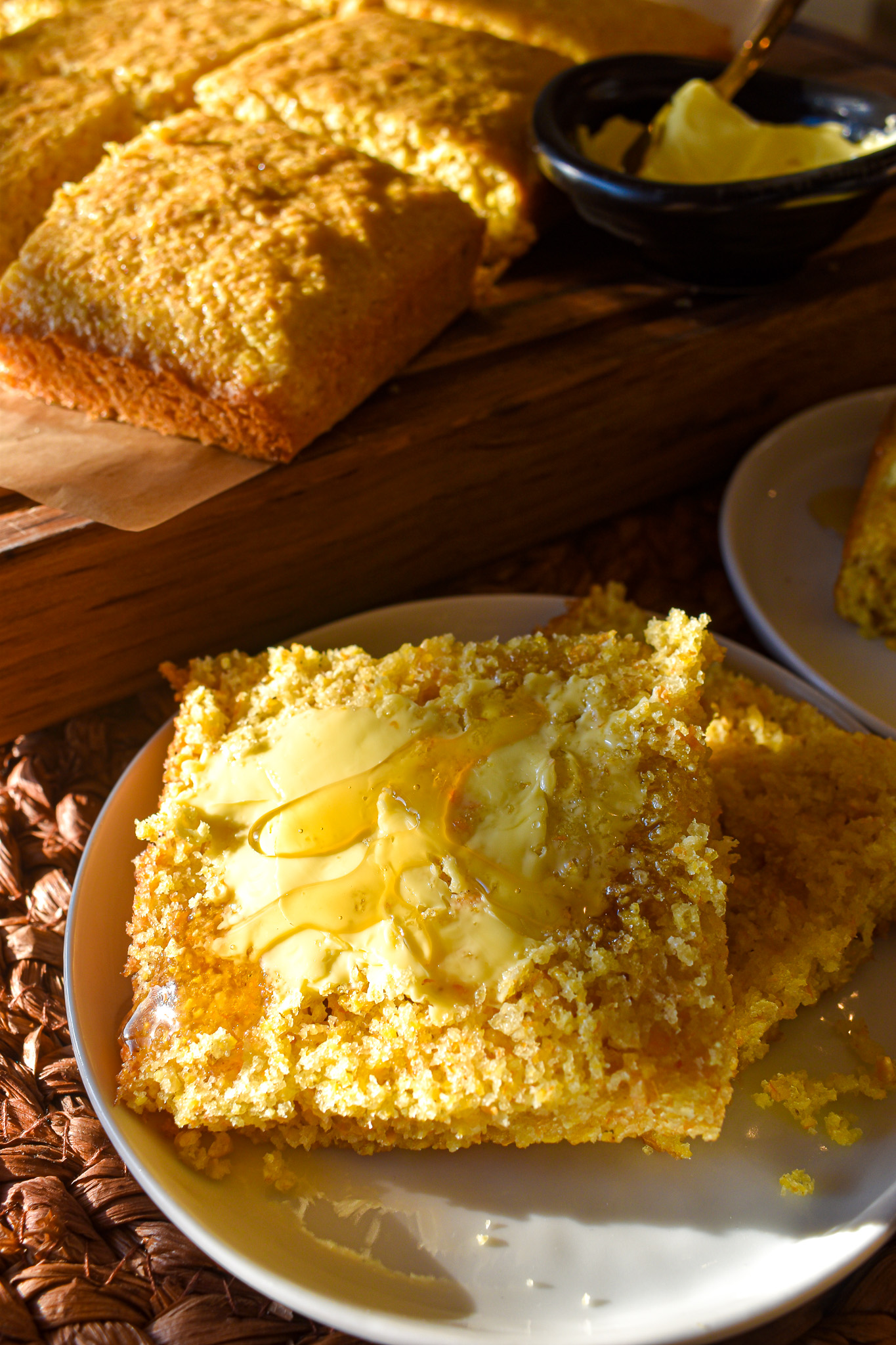 A slice of golden cornbread on a white plate topped with melting butter and a drizzle of honey