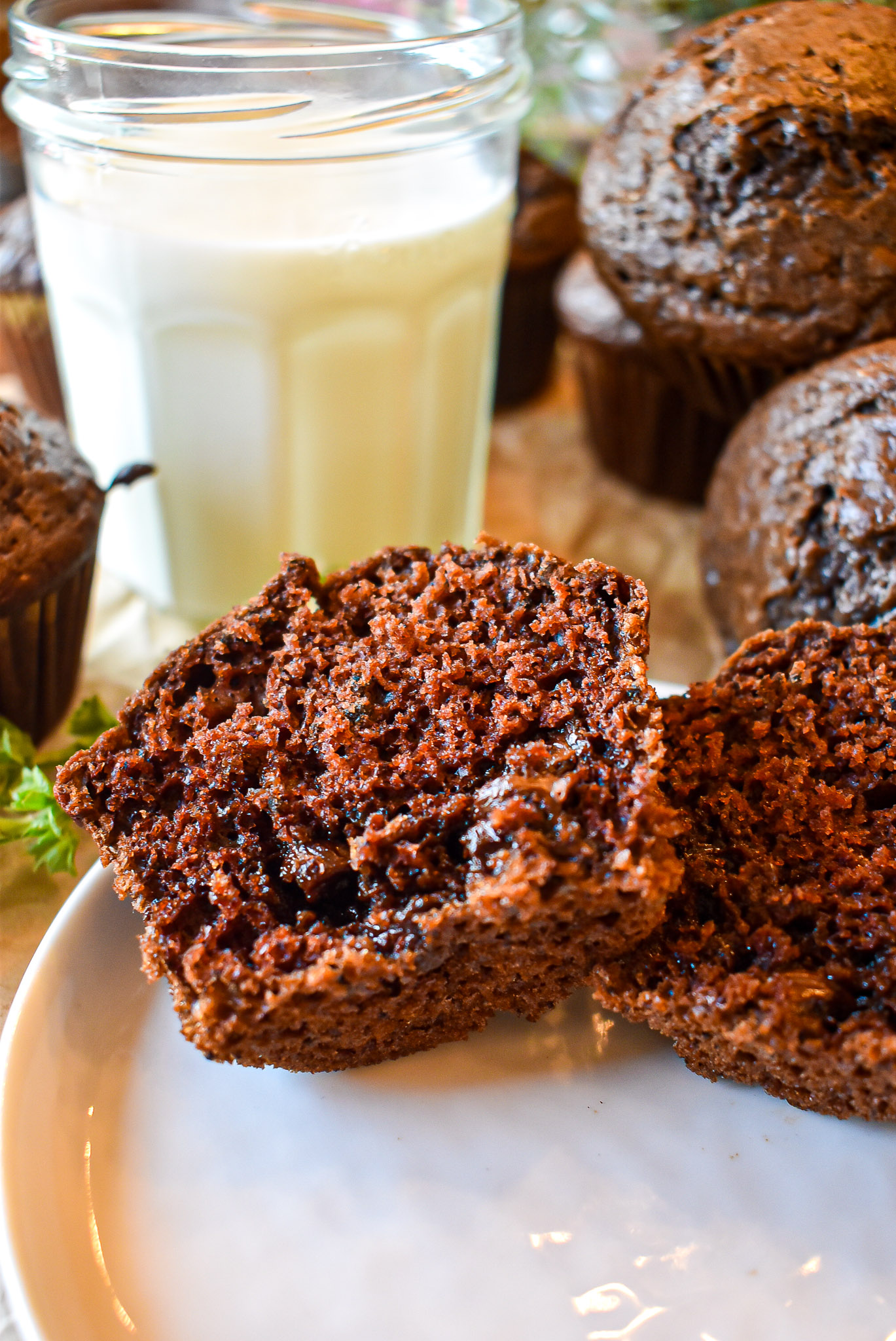 A chocolate muffin split open to show its moist interior, with a glass of milk and whole muffins behind it