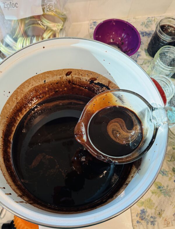 A glass measuring cup holding chocolate milk concentrate scooped from a large white pot